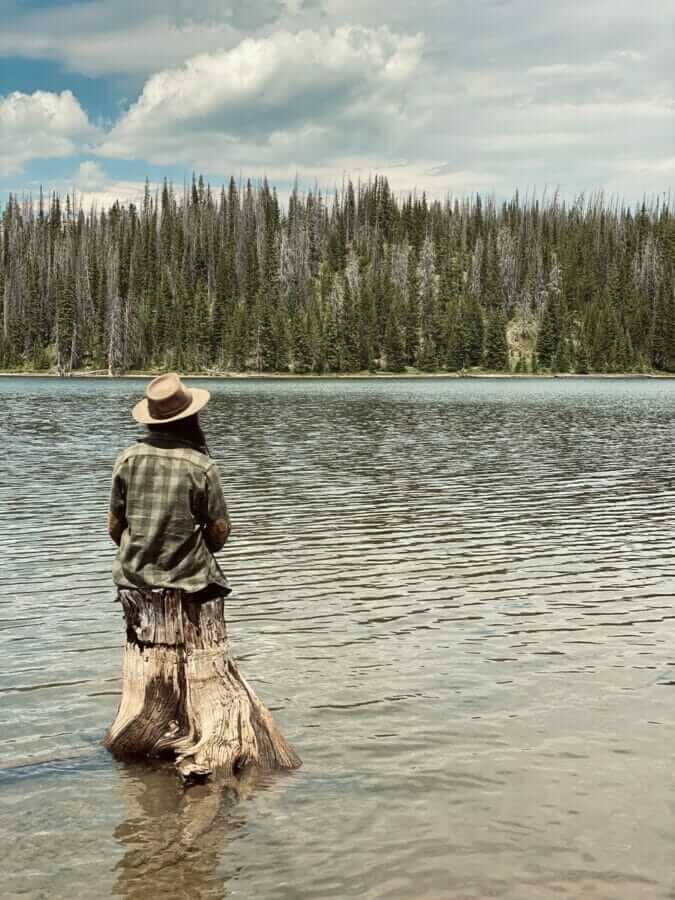 man in brown hat standing on brown wood log on body of water during daytime