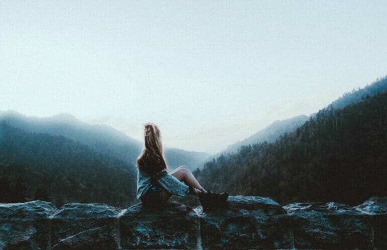 woman sitting on gray rock facing green mountains