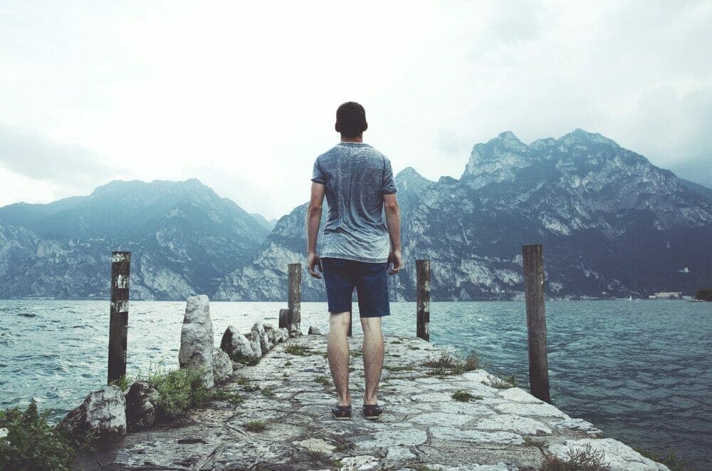 man standing on gray concrete dock facing body of water and mountains at daytime