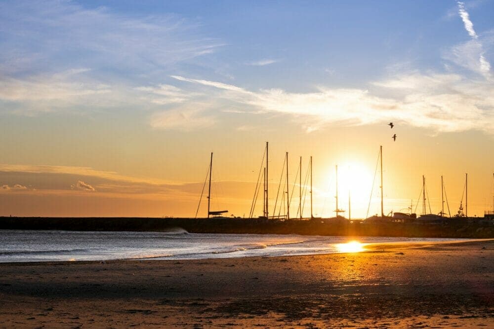 the sun is setting on the beach with sailboats in the background