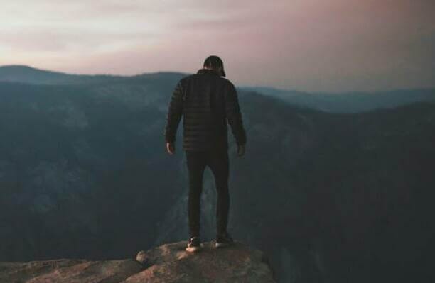 man wearing black bubble jacket on rocky mountain hill in front of mountains under white, pink, and blue cloudy skies during daytime