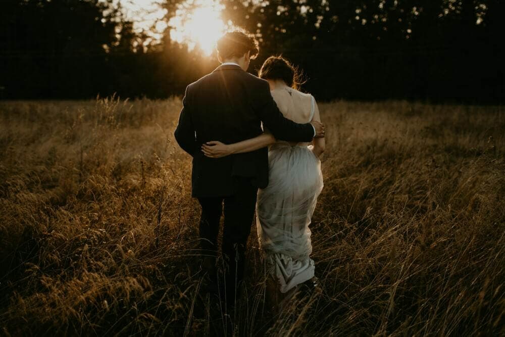 a bride and groom embracing in a field at sunset