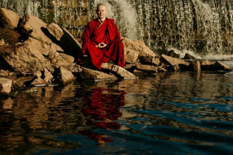 A monk in red robes meditates by a tranquil waterfall, reflecting peace.